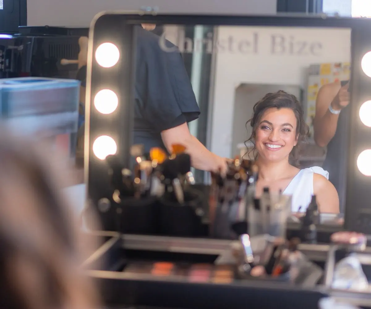 Future mariée découvrant son reflet avec émotion dans le miroir lors de son essai coiffure et maquillage avec la hair & make-up artist.
